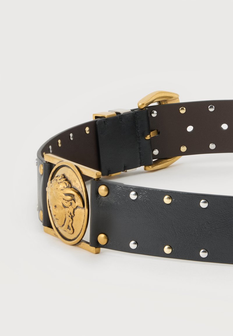 Ceinture en cuir noir avec des clous dorés et argentés et une boucle ovale en or ornée d'une tête de Méduse, présentée sur un fond blanc.