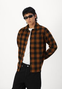 Brun og sort ternet flannel skjorte med knapper, båret over en hvid t-shirt, parret med sorte bukser og solbriller.