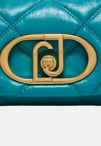 Bolso de piel en color verde azulado con textura acolchada, un cierre de logotipo en tono dorado y herrajes pulidos. Forma ovalada distintiva con acabado brillante.