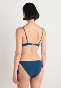 Zils bikini ar regulējamiem siksniņām, zelta detaļām un bezšuvju dizainu. Apakšda�ļa piedāvā vidēju segumu un sānu griezuma detaļu.