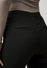 Zwarte, smalle broek met een gladde textuur, voorzien van achterzakken en een elastische tailleband, gedragen met een aansluitend beige top.