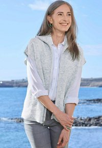 Hellgrauer gestrickter Westen mit hohem Kragen, kurzen Ärmeln und Reißverschluss vorne, über einem weißen Hemd, getragen von einer Frau am Meer.