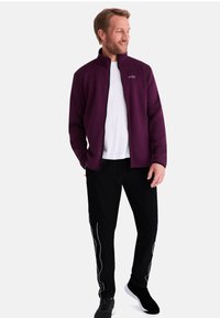 Lila zip-up Jacke über einem weißen T-Shirt, kombiniert mit schwarzen Hosen mit weißen Wellenmustern an den Seiten. Schwarze Sneaker runden das Outfit ab.