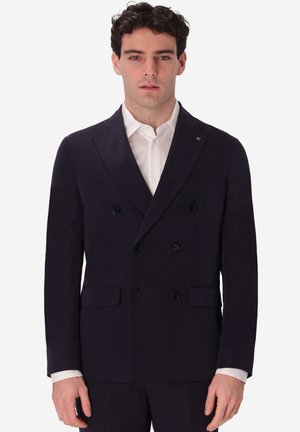 Uomo che indossa un blazer nero doppiopetto sopra una camicia bianca, in piedi davanti a uno sfondo bianco semplice.