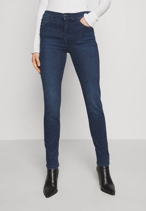 Kvinde iført mørkeblå skinny jeans, hvid ribbet langærmet top og sorte spidse ankelstøvler, stående med den ene hånd i lommen.