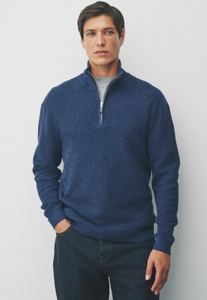 Homme portant un pull bleu marine à col zippé texturé sur une chemise gris clair, les mains dans les poches d'un jean en denim foncé, se tenant contre un mur blanc.