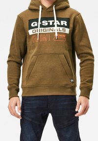 Sudadera G-Star Raw en verde oliva con cordones blancos, gran texto de marca en negro y naranja, bolsillo frontal tipo canguro, dobladillo y puños acanalados.