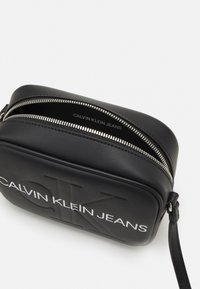 Černá crossbody taška Calvin Klein Jeans se stříbrným zipem a embosovaným logem, vybavená zápěstním popruhem na bílém pozadí.