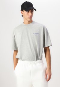 Grå kortermet t-skjorte med rund hals, med blå "ALLSAINTS" tekst. Båret med hvite bukser med elastisk liv og en svart caps.