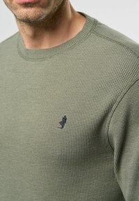 Groene gestructureerde trui met een ronde halslijn en een klein marineblauw geborduurd logo op de linkerkant van de borst. Detail van wafelbreipatroon.
