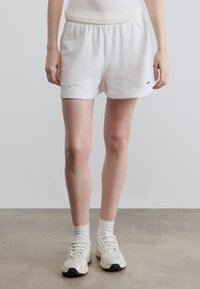 Λευκά βαμβακερά σορτς με ελαστική μέση, διαθέσιμο με πλαϊνές τσέπες και μικρό λογότυπο κοντά στο τελείωμα. Συνδυάζονται με λευκά αθλητικά παπούτσια και κάλτσες.