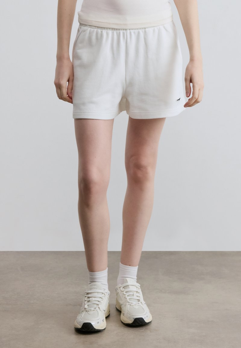 Λευκά βαμβακερά σορτς με ελαστική μέση, διαθέσιμο με πλαϊνές τσέπες και μικρό λογότυπο κοντά στο τελείωμα. Συνδυάζονται με λευκά αθλητικά παπούτσια και κάλτσες.