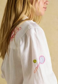 Weißes Hemd aus Leinen mit bunter Stickerei: ein rober Hummer, ein rosa Tennisschläger und ein grüner Tennisball auf den Ärmeln.