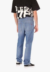 Persona che indossa jeans azzurri chiari, sneakers bianche e nere, e una maglietta nera oversize con lettere bianche in grassetto sulla schiena.