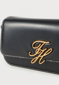 Μαύρο δερμάτινο τσαντάκι χειρός με χρυσή μεταλλική λογότυπο διακόσμηση. Το σχέδιο περιλαμβάνει λευκή ραφή κατά μήκος των άκρων και λεία υφή.