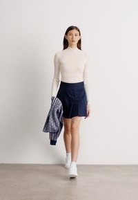 Camisola branca de gola alta com nervuras, saia plissada azul marinho e um casaco estampado. Modelo usa ténis brancos, em pé sobre um chão de cor clara.