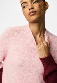 Svijetlo ružičasti pleteni pulover s V-izrezom i rebrastim bordo rukavima. Tekstura je meka i pahuljasta, s suptilnim mrljastim uzorkom.