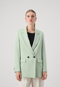 Blazer de color verde claro, de doble botonadura, con solapas muesca, botones negros y dos bolsillos frontales, llevado sobre una blusa negra. Textura de tela suave.