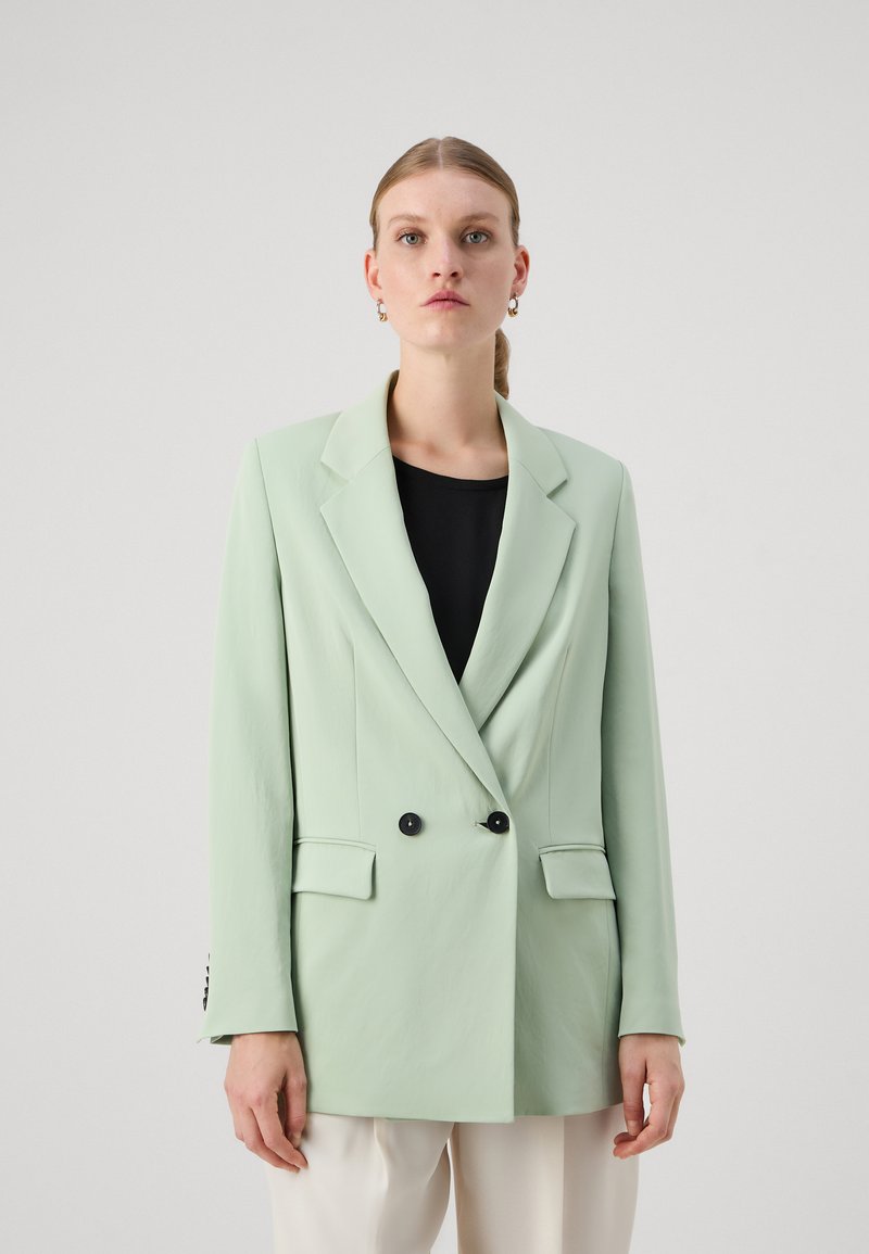 Blazer de color verde claro, de doble botonadura, con solapas muesca, botones negros y dos bolsillos frontales, llevado sobre una blusa negra. Textura de tela suave.