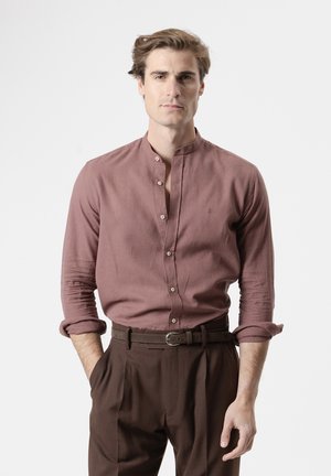 Hombre de pie mirando hacia adelante, con una camisa color malva con cuello mandarín y mangas remangadas, y pantalones de tiro alto marrón oscuro con cinturón.
