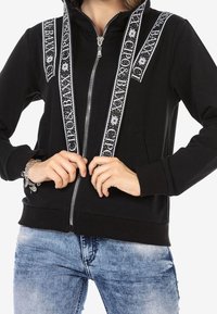 Black zip-up hoodie with white patterned trim along the shoulders and sides. Features a front zipper and two side pockets.