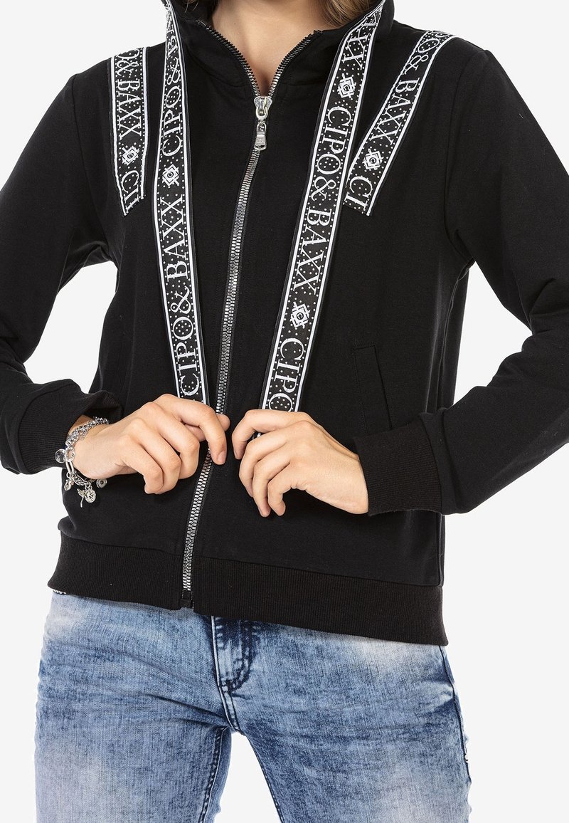 Black zip-up hoodie with white patterned trim along the shoulders and sides. Features a front zipper and two side pockets.