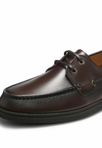 Chaussure bateau en cuir marron foncé avec lacets noirs, œillets métalliques, coutures visibles et semelle en caoutchouc noire.