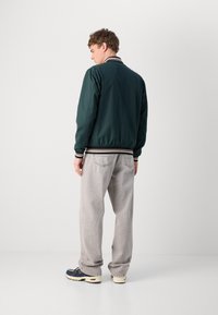 Chaqueta bomber verde con cuello y puños de rayas acanaladas, combinada con pantalones grises claros y zapatillas, vista trasera.