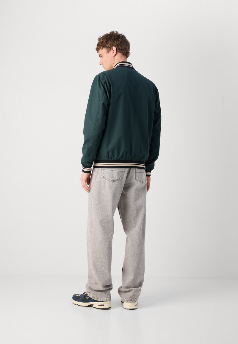 Veste bomber verte avec col et poignets rayés côtelés, associée à un pantalon gris clair et des baskets, vue de dos.