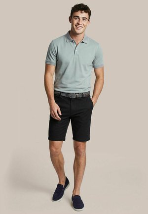 Jonge man staat en lacht, draagt een lichtgrijs poloshirt, zwarte korte broek met een patroonriem en marineblauwe instapschoenen.