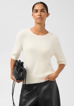 Femme portant un pull blanc duveteux à manches mi-longues et un pantalon en cuir noir, tenant un petit sac à main en cuir noir avec une fermeture à cordon.