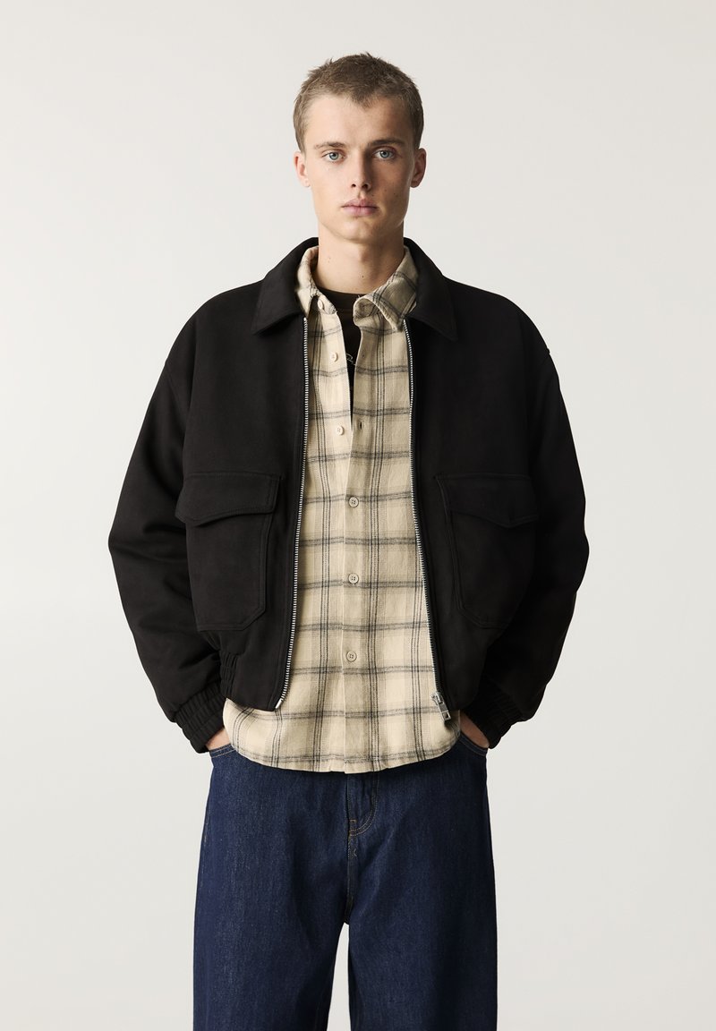 Svart bomberjacka med två framfickor, bärs över en beige rutiga skjorta. Modellen har mörkblå denimjeans till. Slät textur, avslappnad design.