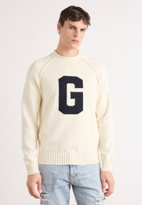 Cream knit sweater with a round neck, ribbed cuffs and hem, featuring a large navy letter "G" on the front. Worn with light blue distressed jeans.