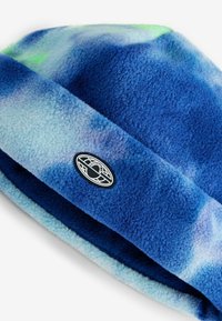Blå fleece hue med en blanding af lyse og mørke blå nuancer. Har et rundt sort patch-logo på folden. Blødt og tekstureret materiale.