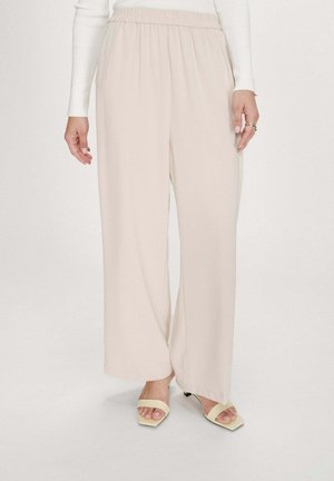 Femme portant un pantalon ample couleur crème, un haut blanc à manches longues, et des sandales à talons ouvertes beige, debout sur un fond blanc uni.