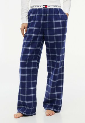 Persona con pantalones de pijama a cuadros azules y blancos con cintura elástica y pies descalzos, de pie sobre un fondo blanco simple.