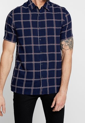 Mężczyzna w krótkim rękawie granatowej koszuli w biało-czerwoną kratę, w czarnych spodniach, z widocznym tatuażem na ramieniu.