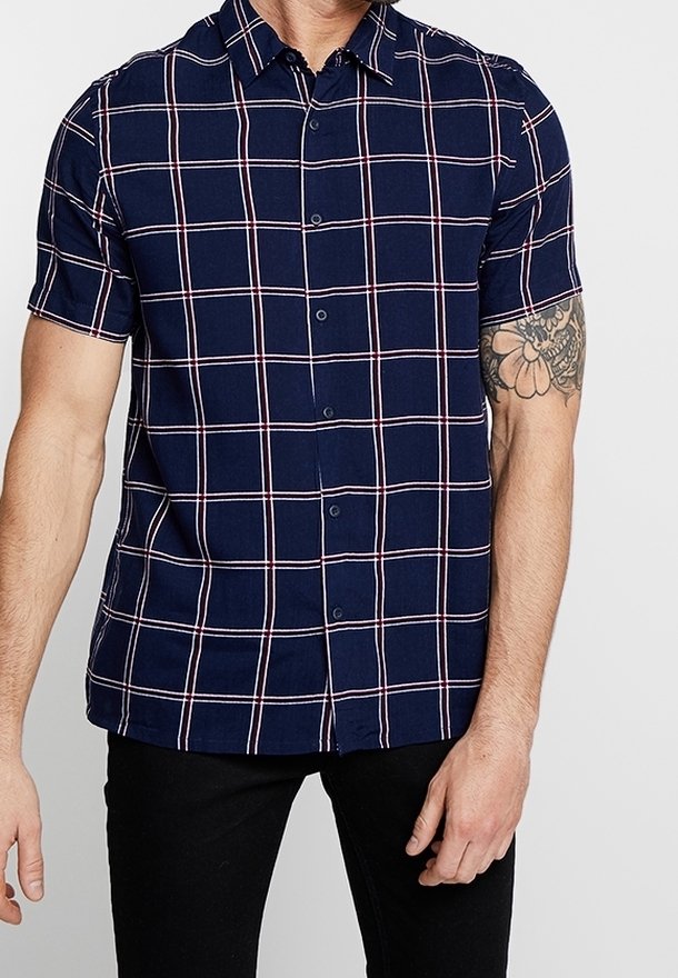 Homme portant une chemise à manches courtes bleu marine à motif à carreaux blanc et rouge, assortie à un pantalon noir et avec un tatouage visible sur le bras.