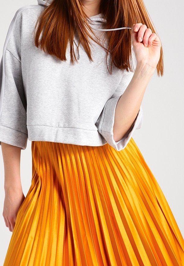 Persona vestida con una sudadera recortada de color gris claro con mangas remangadas y una falda plisada de un brillante color naranja, sosteniendo un mechón de cabello.