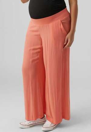 Person trägt weite Korallenhose, schwarzes Oberteil und weiße Sneaker, steht vor einem schlichten grauen Hintergrund.