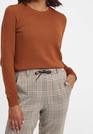 Mujer con suéter de manga larga texturizado color óxido y pantalones de cuadros de talle alto con cordón negro, con una mano en el bolsillo.