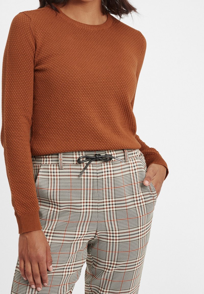 Femme portant un pull à manches longues texturé orange rouille et un pantalon à carreaux taille haute avec un cordon noir, une main dans la poche.