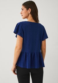 Bluse aus blauer Spitze mit kurzen fransigen Ärmeln, floralem Muster, einem Peplum-Saum und kleinen Ausschnitten. Getragen über schwarzen, figurbetonten Hosen.