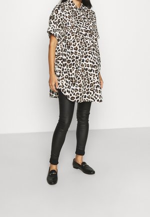 Femme portant une chemise ample à manches courtes imprimée léopard, un pantalon noir ajusté, et des mocassins noirs, debout sur un fond blanc uni.