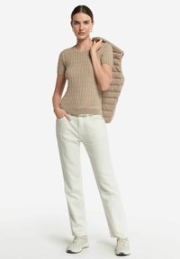 Beige gerippter Strick-Top mit kurzen Ärmeln, kombiniert mit weißen gerade geschnittenen Hosen und hellen Sneakers. Das Model hält eine hellbraune Steppweste auf einem Arm.