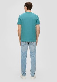 Een teal kortemouwen t-shirt gezien vanaf de achterkant, gecombineerd met lichtblauwe jeans en witte sneakers. Eenvoudig ontwerp, zachte textuur.