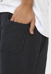 Pantalones negros texturizados con una mano en el bolsillo. Presentan un ajuste holgado y un sutil detalle en la tela que añade interés visual.