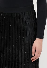 Sort plisseret fløjlsskjørt med en bred talje; prydet med små, skinnende udsmykninger for detalje; blød tekstur og lodrette linjer.