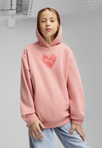 Sudadera rosa de gran tamaño hecha de una tela suave, con un logo en forma de llama en un rosa más claro. Manga larga con puños acanalados, combinada con jeans de color azul claro.