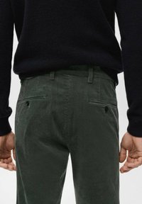 Mężczyzna noszący ciemnozielone spodnie sztruksowe z zapinanymi na guziki tylnymi kieszeniami oraz czarny sweter, widoczny od pasa w dół, widok od tyłu.